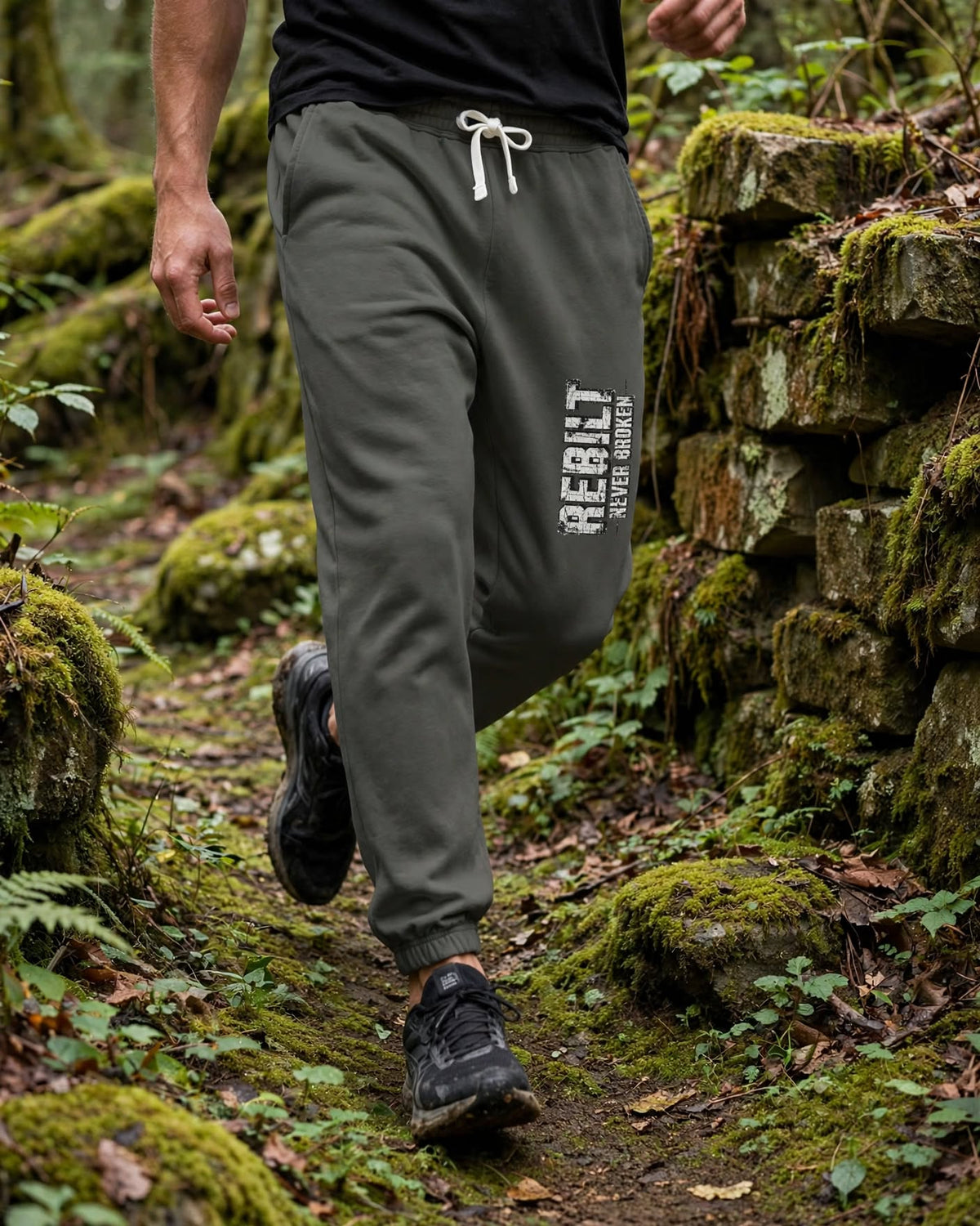 Man walking a mossy forest trail in Pepper REBILT Never Broken joggers, vertical distressed white REBILT print on the left thigh