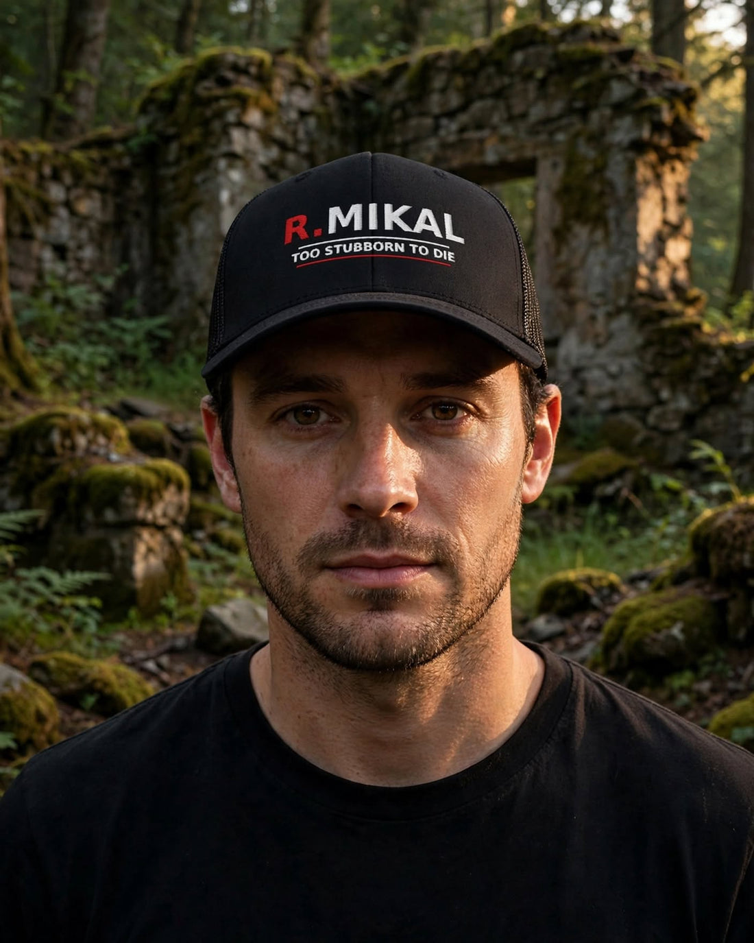 Man in forest ruins wearing the black R.Mikal Too Stubborn to Die trucker hat, red R. and white MIKAL above TOO STUBBORN TO DIE