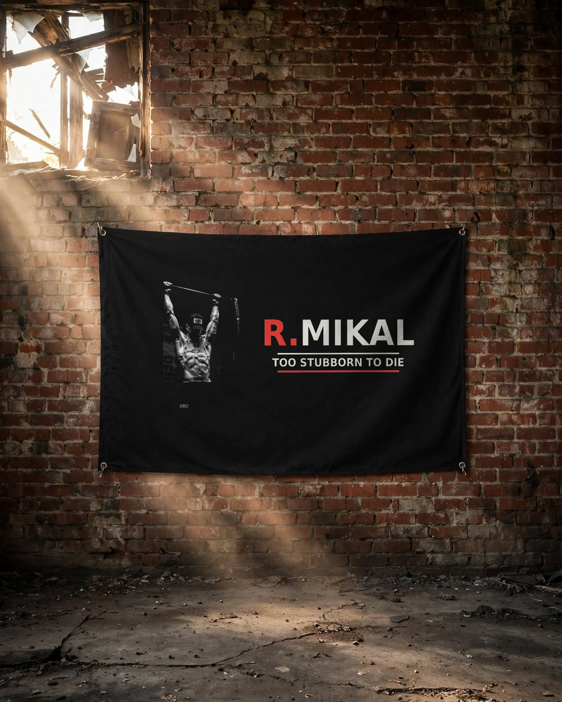 R.MIKAL Too Stubborn to Die signature flag hung on a brick wall in an old warehouse with light through a broken window