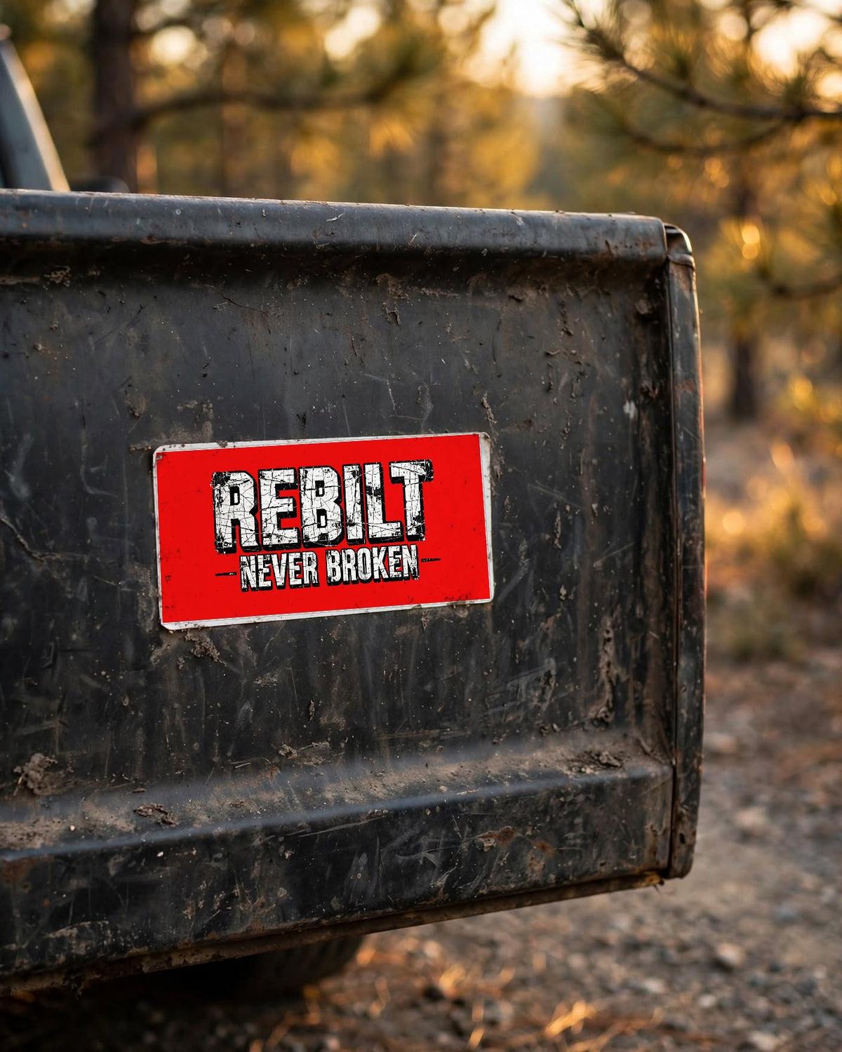 Red REBILT Never Broken bumper sticker on the dusty tailgate of an old pickup truck in a pine forest